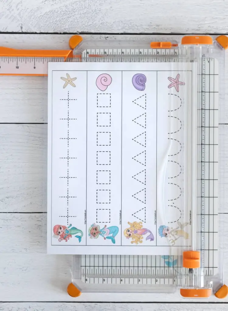 A picture of a page of mermaid themed prewriting practice cards on a paper trimmer. The page has four strips that are being cut along a solid line to form prewriting practice cards for preschoolers. 