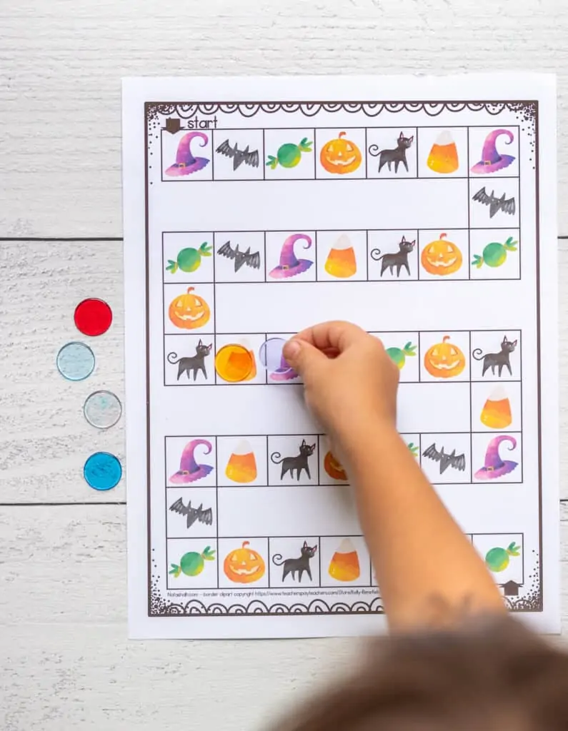 A child's hand placing a plastic chip on a printable board game with Halloween images. 