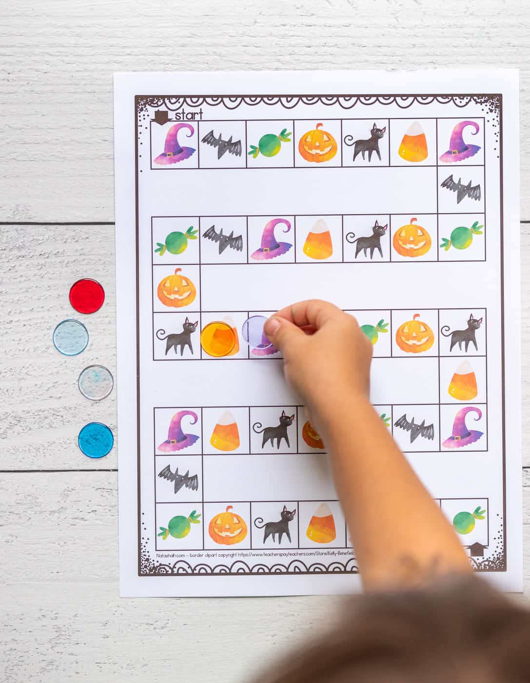 A child's hand placing a plastic chip on a printable board game with Halloween images.
