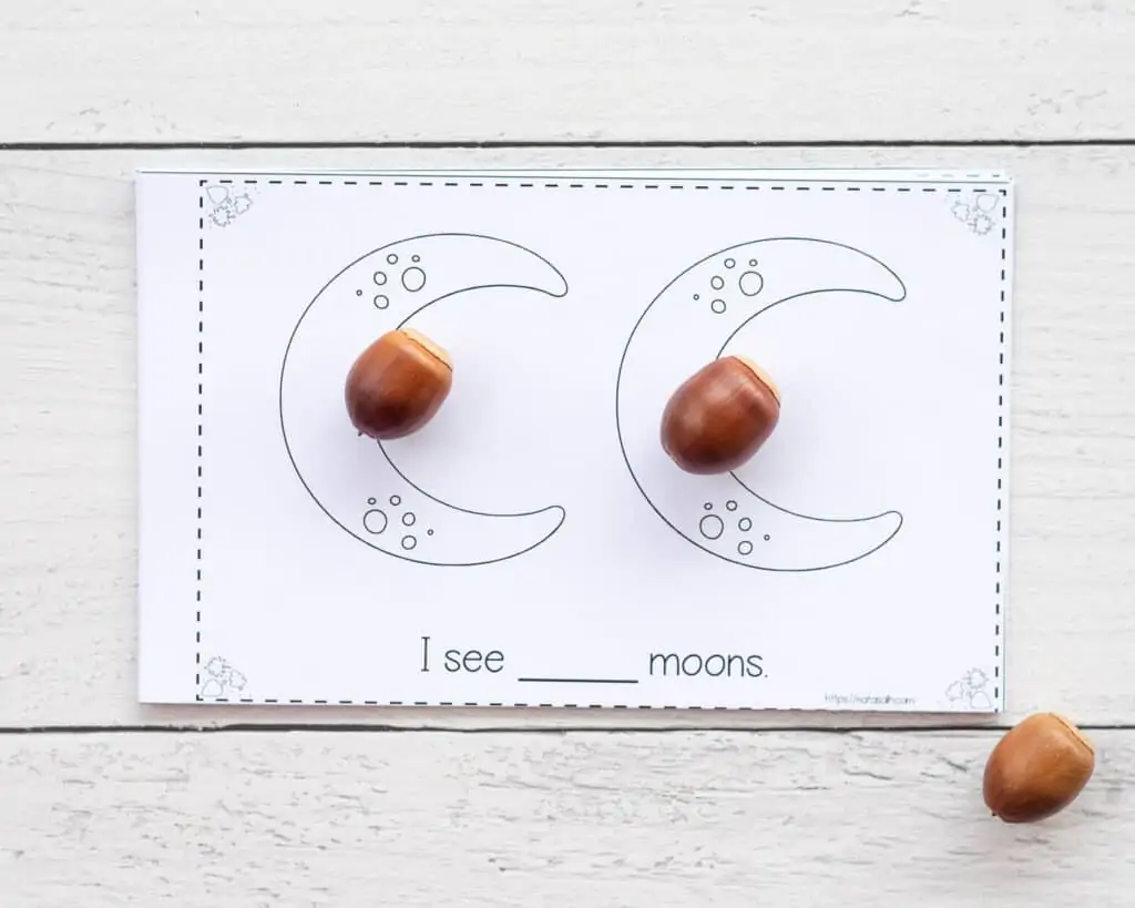 A printed page with two moons and the text "I see ___ moons" below. There is an acorn on top of each moon.