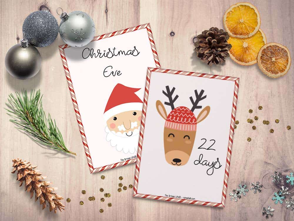 A preview of two 5x7 Advent calendar cars for kids. One has "22 days" with a reindeer and the card behind has "Christmas Eve" with a Santa face. The cards are on a light wood grain background with assorted Christmas items like ornaments, glitter, and orange slices.