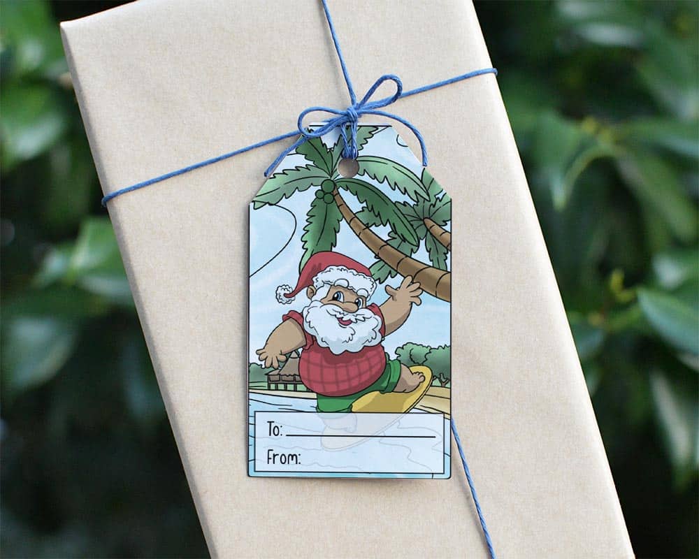 A preview of a printable tropical Christmas gift tag featuring a tanned, surfing Santa at the beach. The tag is tied to a brown paper wrapped package with blue twine.