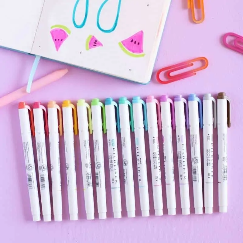 A set of colorful Zebra midline markers on a pink background with a notebook and three colorful plastic paperclips.