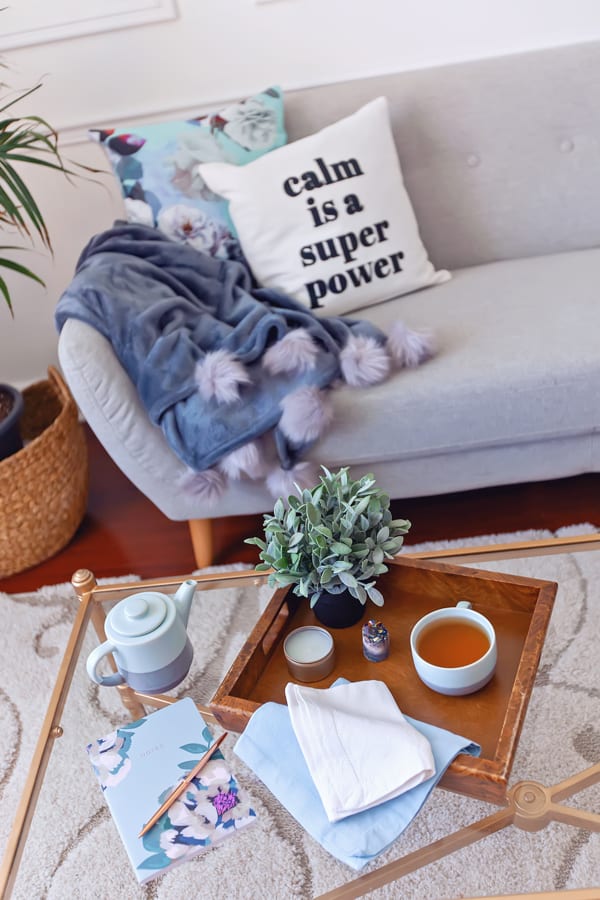 An image of a coffee table in front of a sofa. The sofa has a throw pillow with "calm is a super power' on it. The table has a notebook and a tea tray with tea, towels, a candle, a crystal, and a small plant on it.