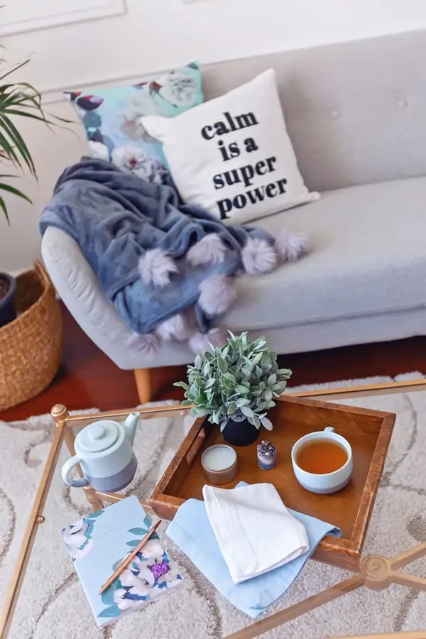 An image of a coffee table in front of a sofa. The sofa has a throw pillow with "calm is a super power' on it. The table has a notebook and a tea tray with tea, towels, a candle, a crystal, and a small plant on it.