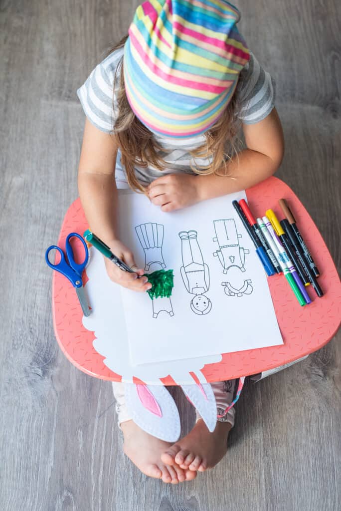 A young girl coloring a paper doll coloring page. The page is resent on a pink lap desk and she is using a green marker to color in a dress.