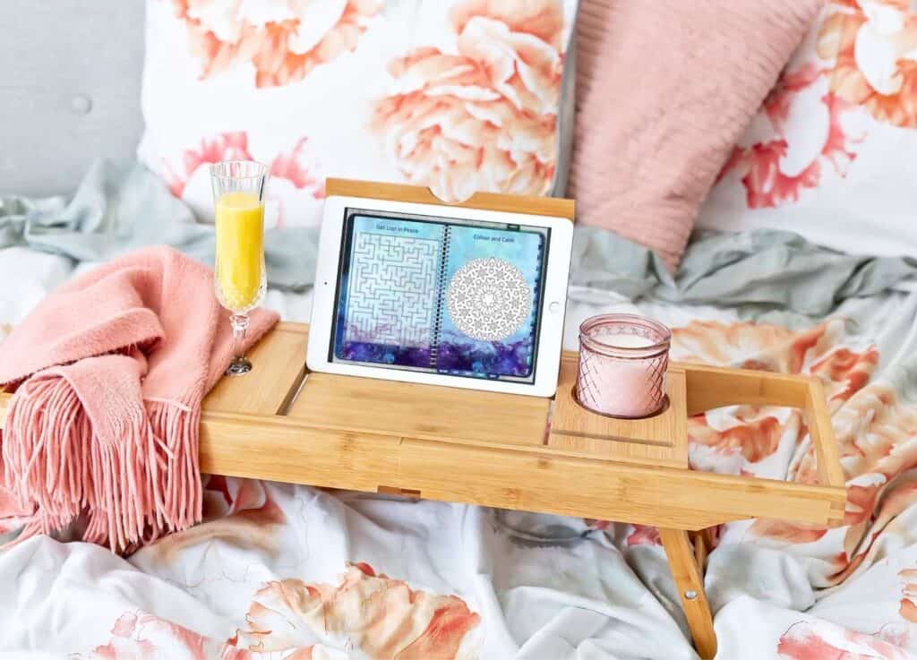 A wood lap desk with a pink candle, blanket, a glass or orange juice, and a white iPad open to a digital planner page with a maze and a mandala to color.