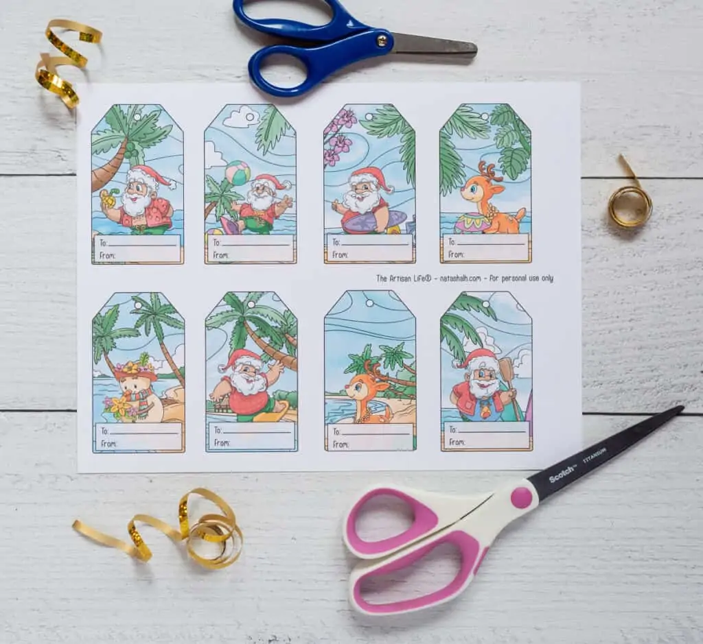 A printed sheet with four tropical Santa gift tags on a white wood surface with two pairs of scissors and curls of gold ribbon