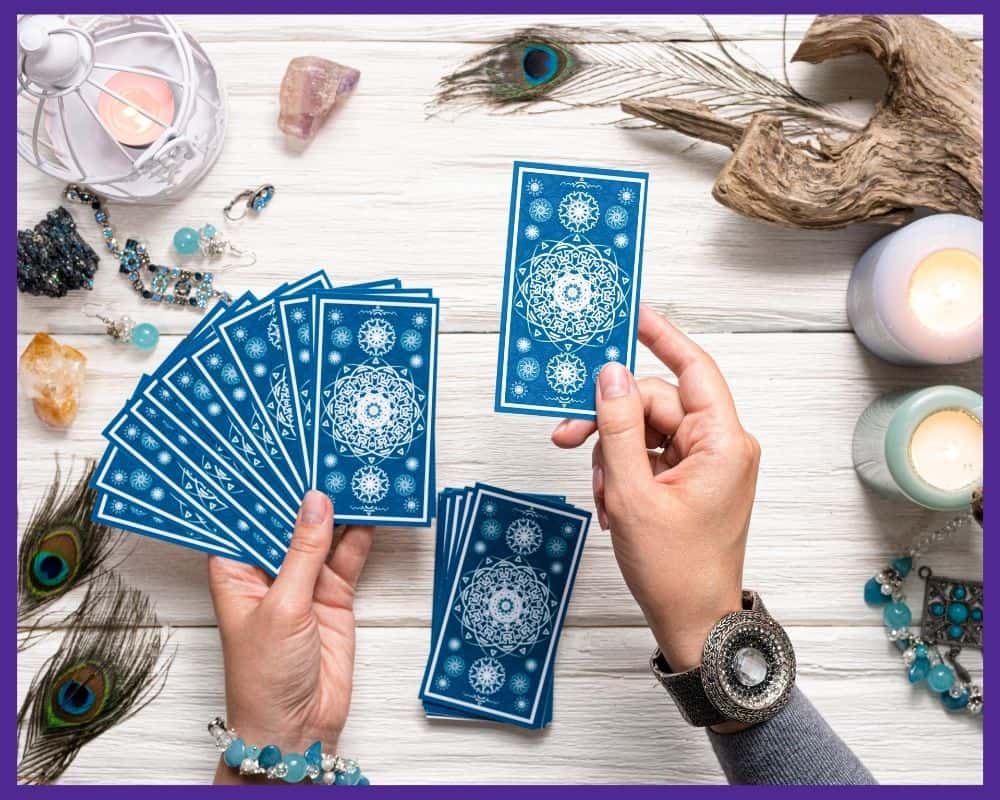 An image a woman's hands holding a blue backed tarot card in her right hand and a fan of cards in her left hand. All cards are face down.