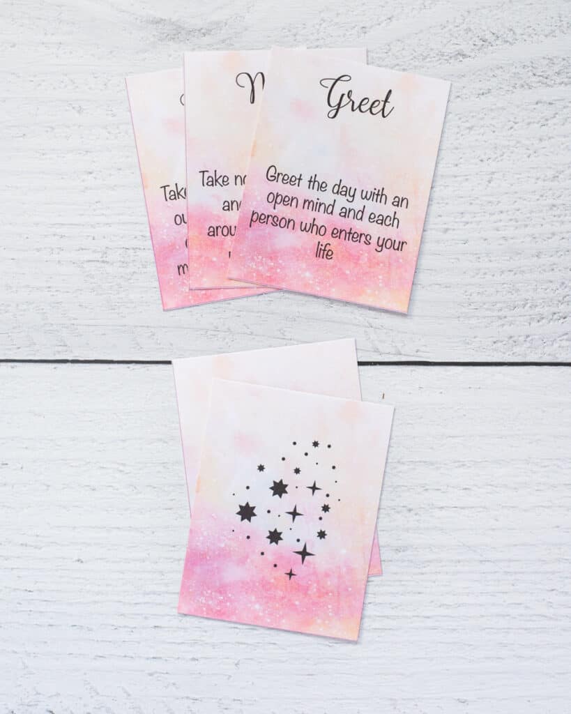 A preview of printed word of the day oracle cards. On top are there cards and below are the back sides of two cards with a pink watercolor background and black stars.