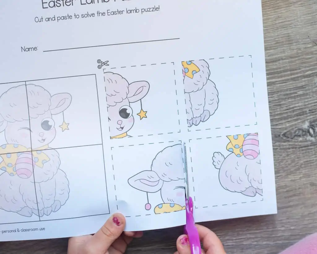 A top down view of a child's hands using pink scissors to cut the pieces from a four put cut and paste puzzle with an Easter lamb.
