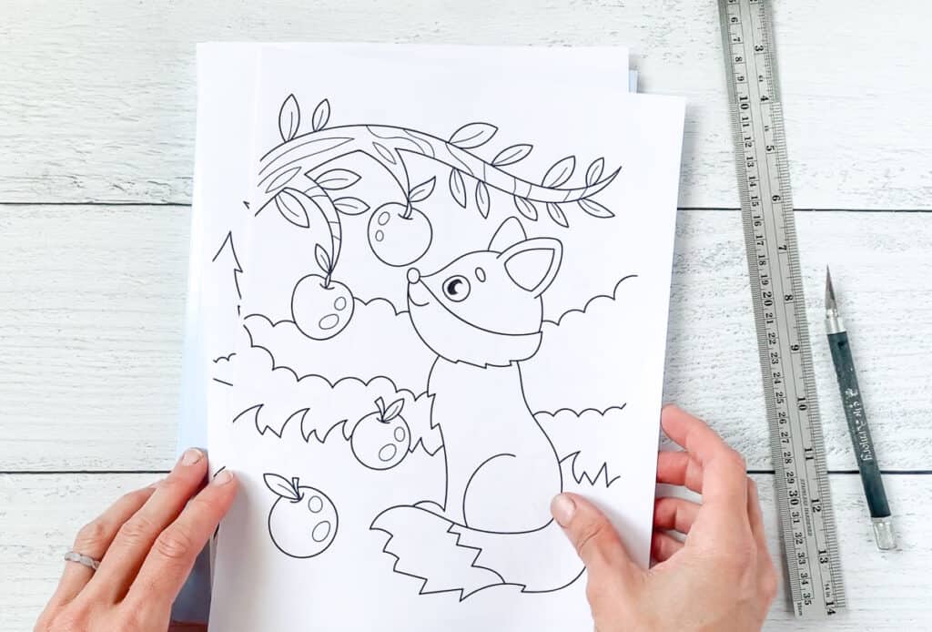 Hands holding cut out coloring page with a fox looking at an apple tree