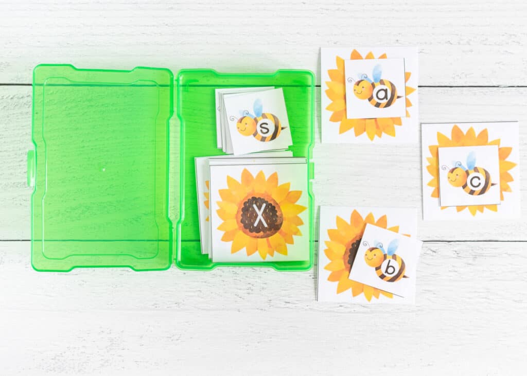 Cards from a bee themed printable alphabet matching game on a white wood surface next to a small green plastic box with additional cards