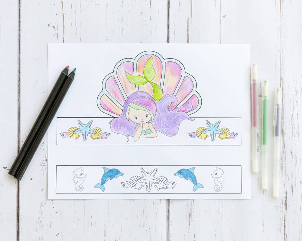 A partially colored printable crown featuring a mermaid with a shell behind her and multiple smaller shells beside her. The crown is shown with two multicolor pencils and three gel pens.