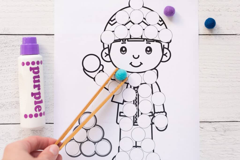 A hand using a pair of bamboo toast tongs to place a blue pompom on a dot marker page
