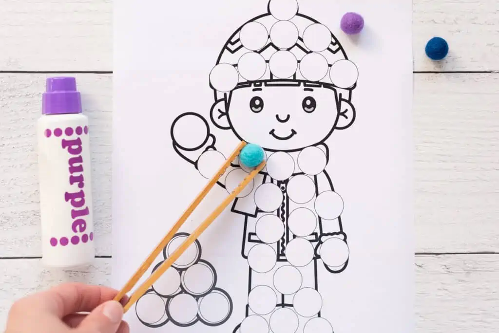 A hand using a pair of bamboo toast tongs to place a blue pompom on a dot marker page