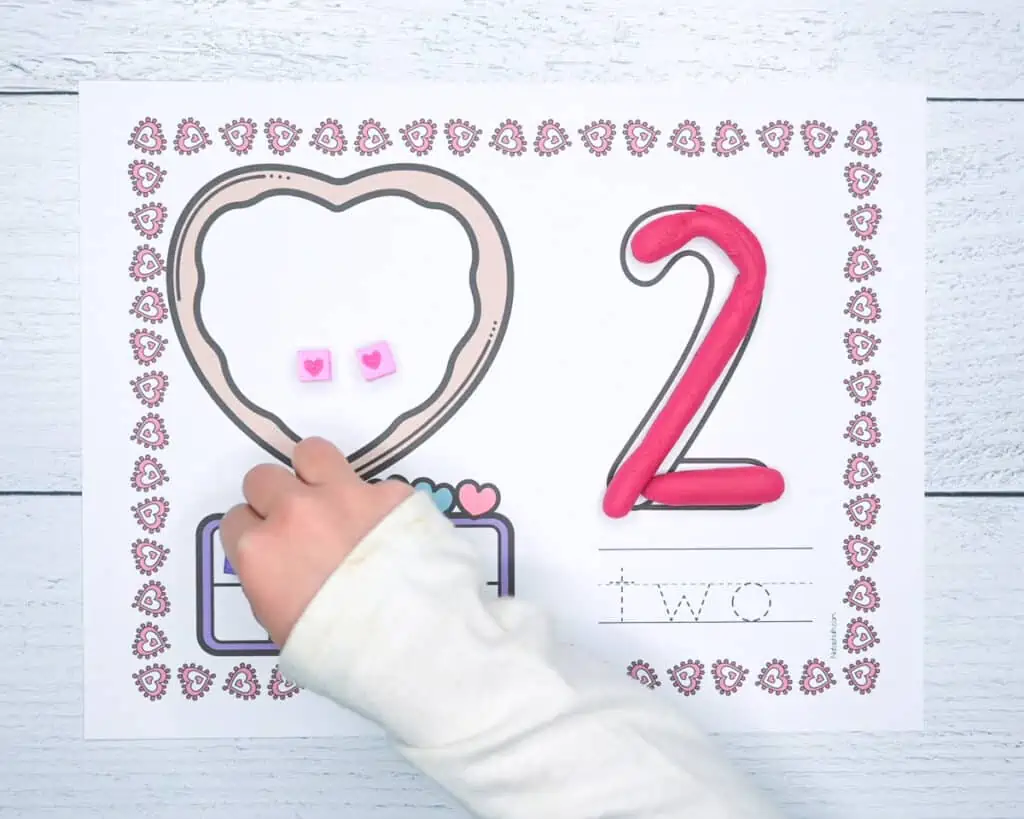 A child's hand placing a bead on a ten frame on a counting mat with a Valentine's Day theme.