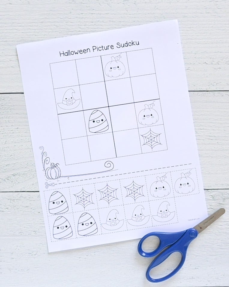 A top down picture of a printed out Halloween picture sudoku game with a 4x4 grid. It is shown on a white wood background with a pair of children's scissors.