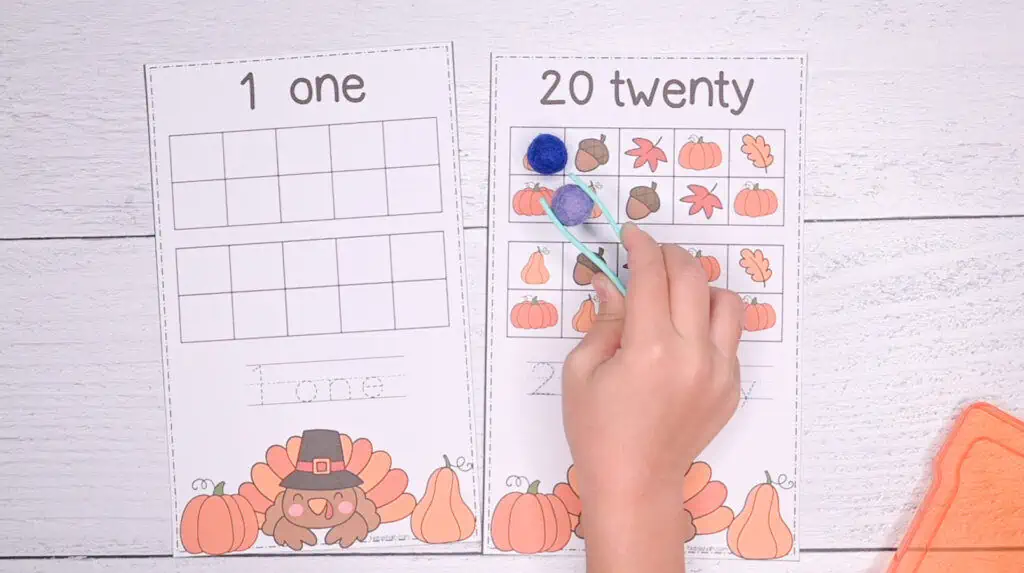 A preview of two double ten frame cards. One shows 1 and the other shows 20. A child is placing a pom pom on the 20 frame with a pair of plastic tweezers.