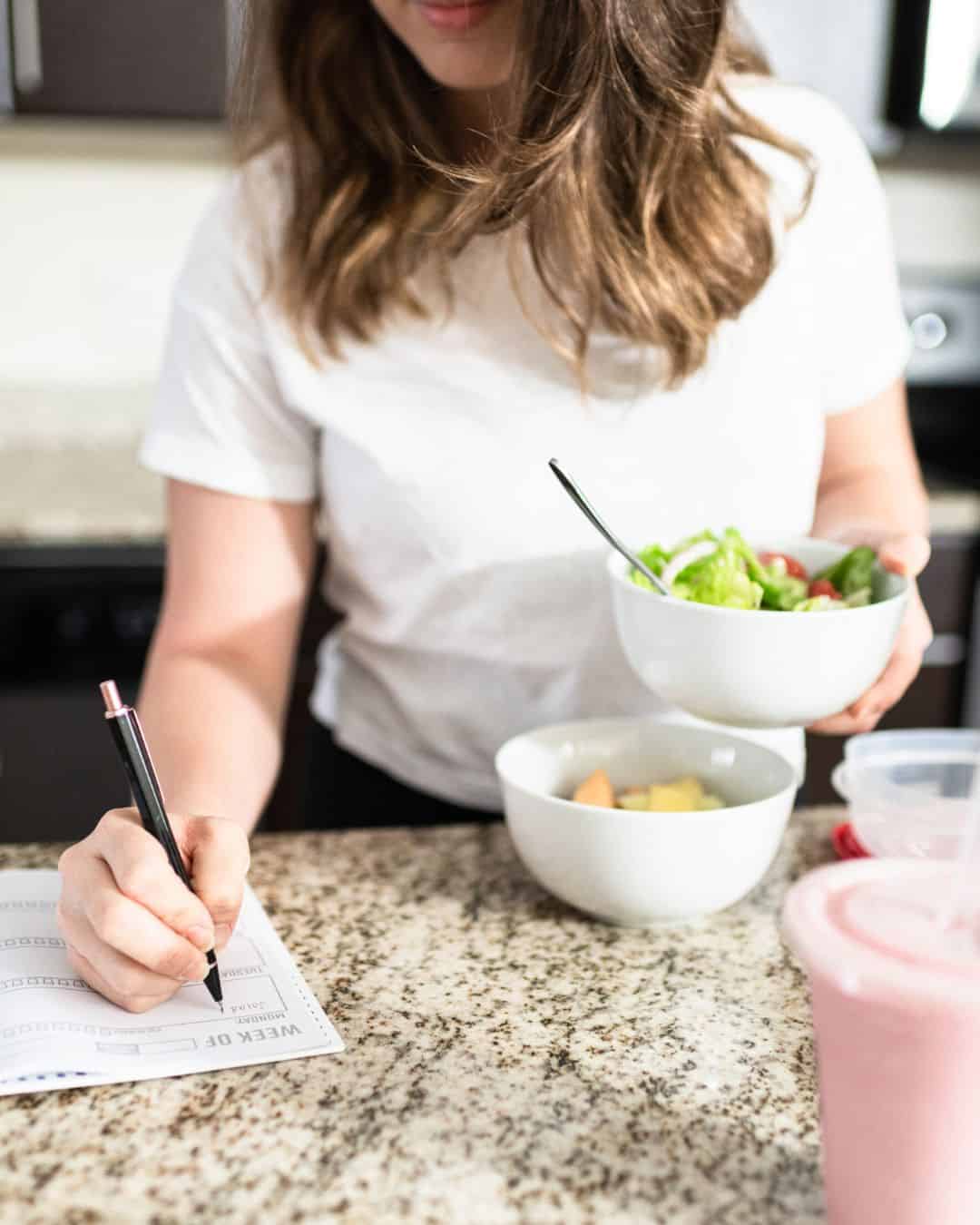 A photo of a woman writing in a planner while standing at her kitchen counter with a bowl of salad