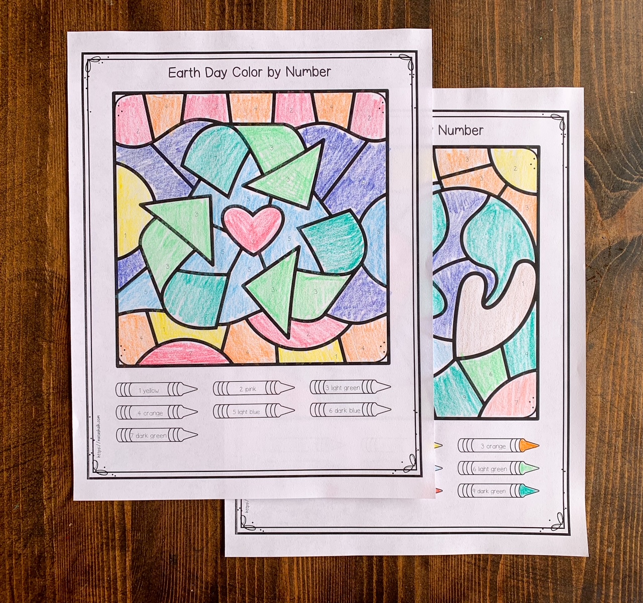 A photograph of two color by number worksheets with an Earth Day theme. One has hands holding a globe and the other has a recycle symbol 
