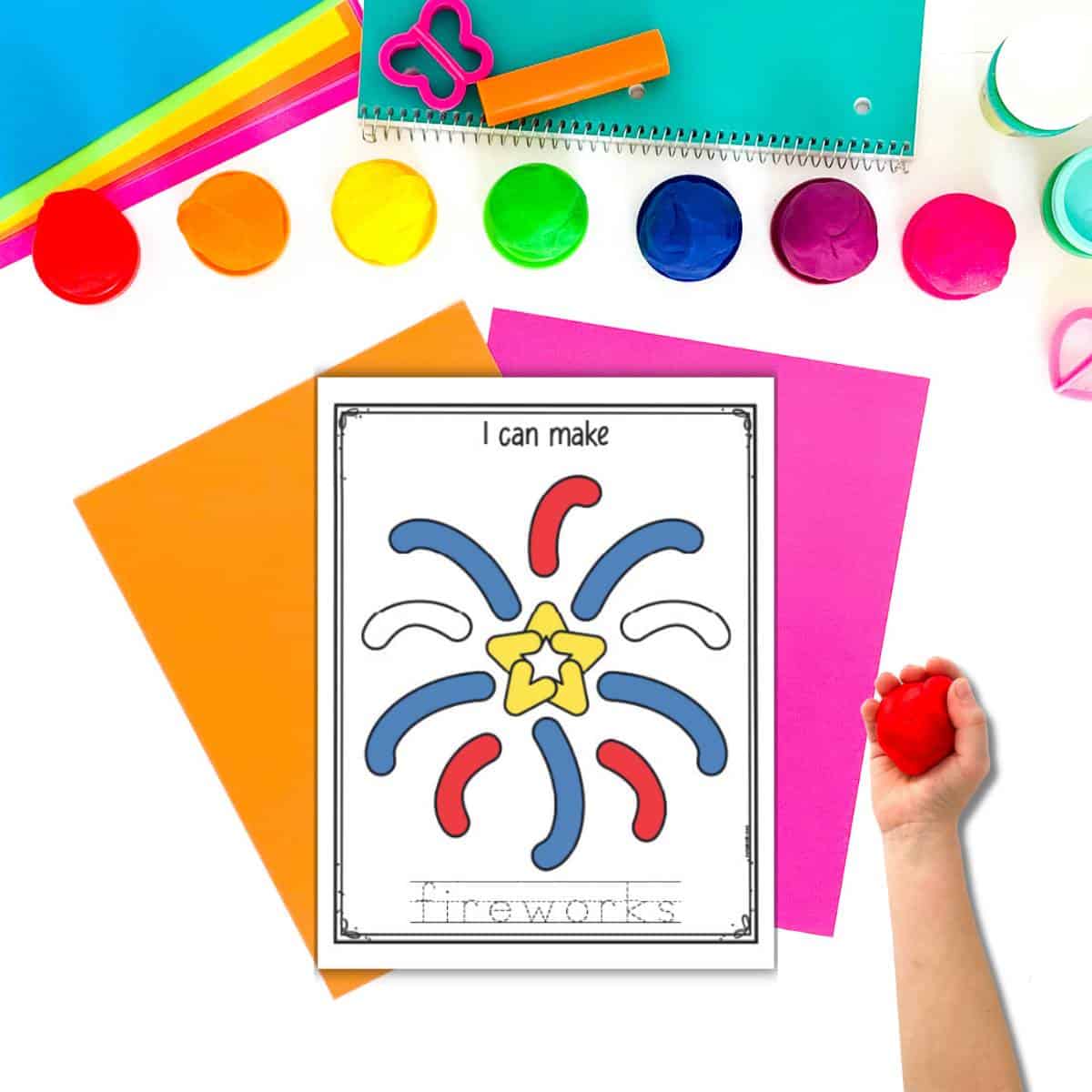 A top down image of paper and play dough with a child's hand holding a ball of red play dough. A play dough activity mat with fireworks is on top of other brightly colored pieces of paper.