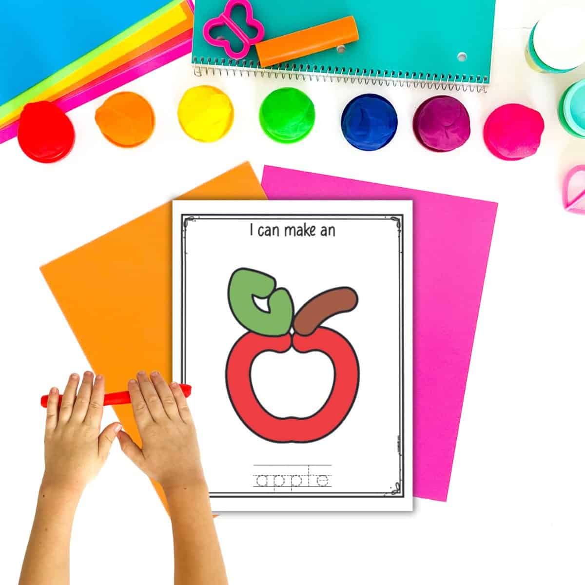 A top down image of paper and play dough with a child's hand rolling a snake of red play dough. A play dough activity mat with an apple is on top of other brightly colored pieces of paper.