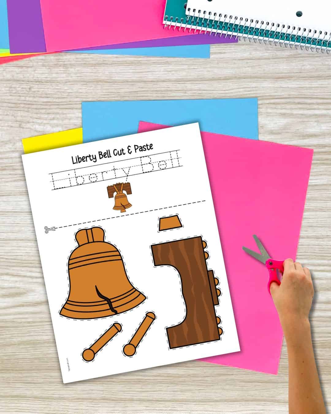 A Liberty Bell cut and paste craft with five pieces on the page. There is also space to trace the words "Liberty Bell" and a child's hand with a pair of blunt scissors.