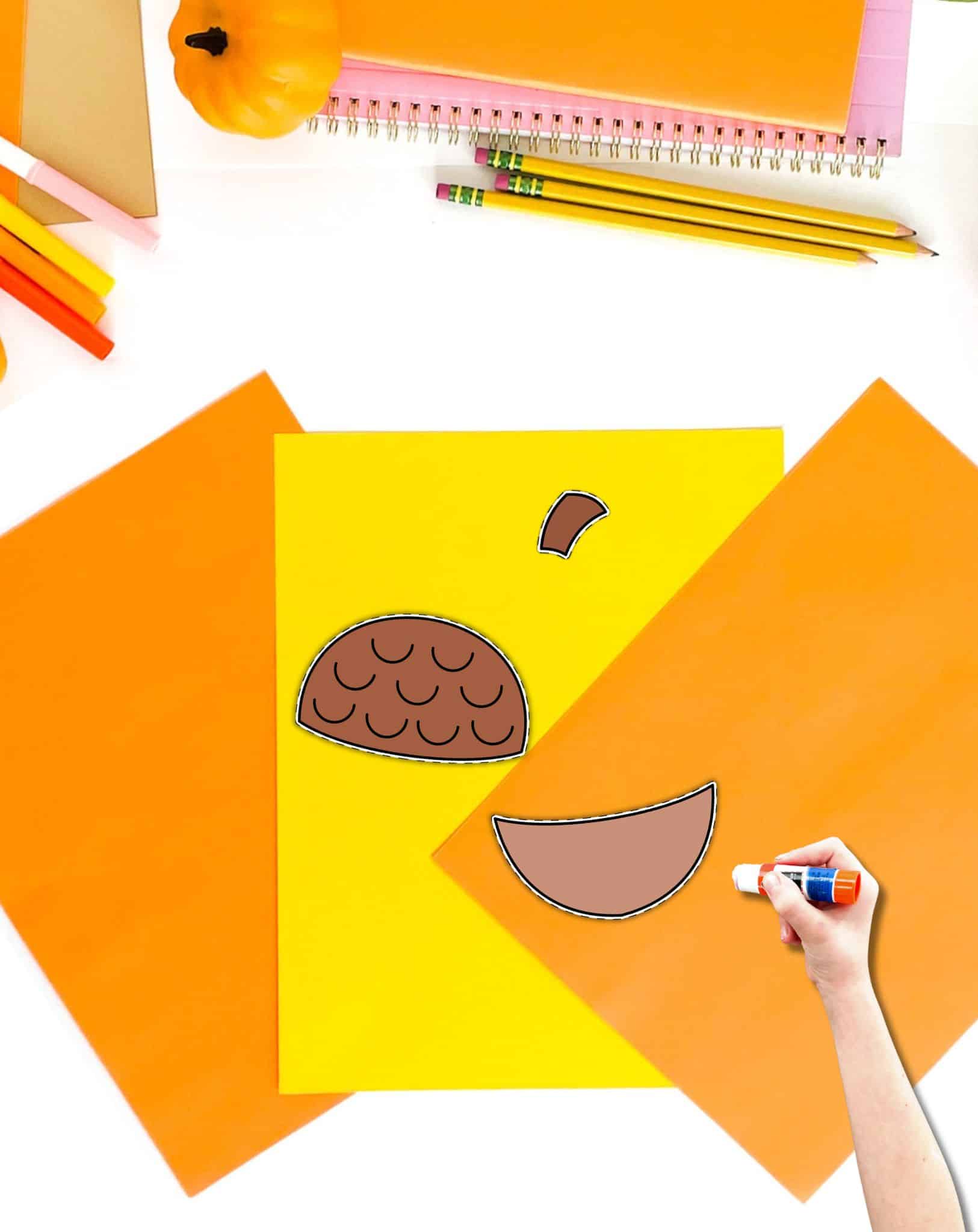 Three pieces of cut and paste acorn craft on a desk with fall school supplies. A child's hand holding a glue stick is also in the frame.