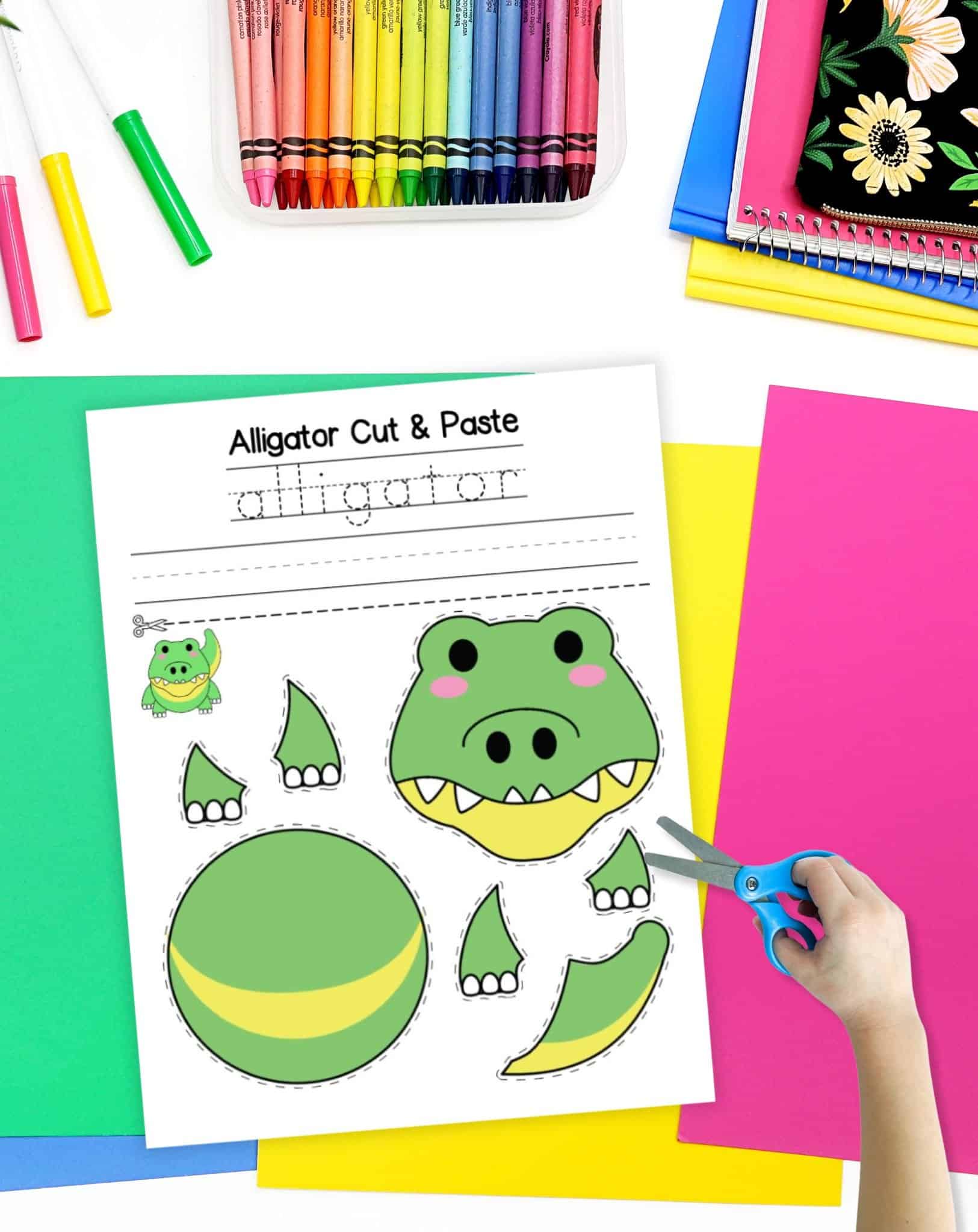 A top down view of a printable cut and paste alligator craft on a desk with colorful papers and crayons. A child's hand with scissors is visible in the foreground.