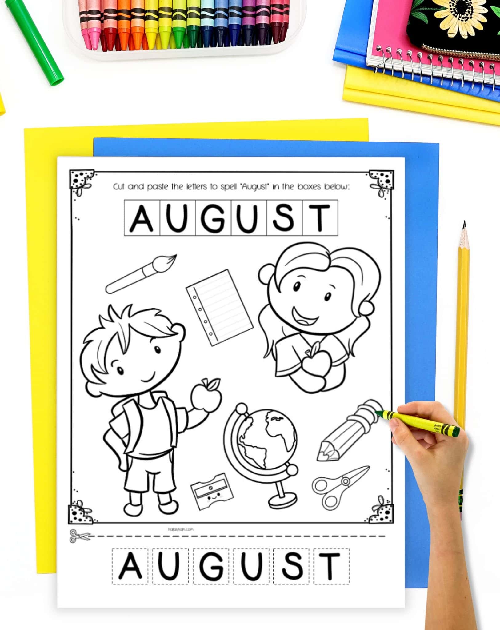 A top down image of an August coloring page and cut & paste worksheet. It is on a desk with colorful school supplies and a hand with a glue stick