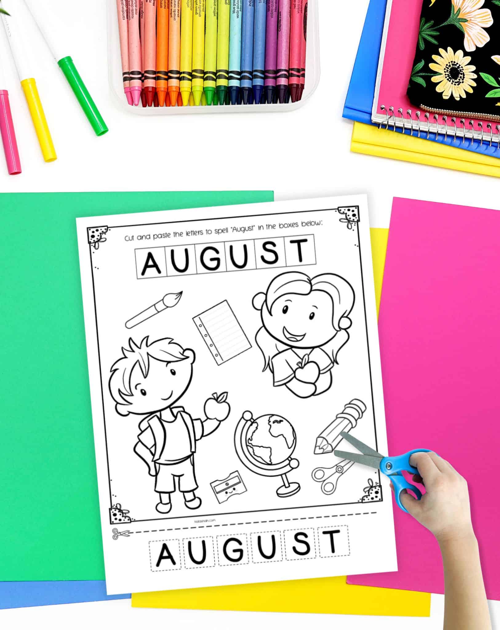 A top down image of an August coloring page and cut & paste worksheet. It is on a desk with colorful school supplies and a hand holding a pair of scissors. 