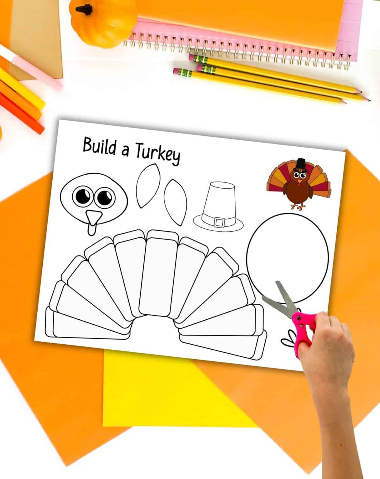 A preview of a printed build a turkey craft page with a child's hand with scissors. The page is shown on top of orange and yellow paper with fall colored craft supplies.