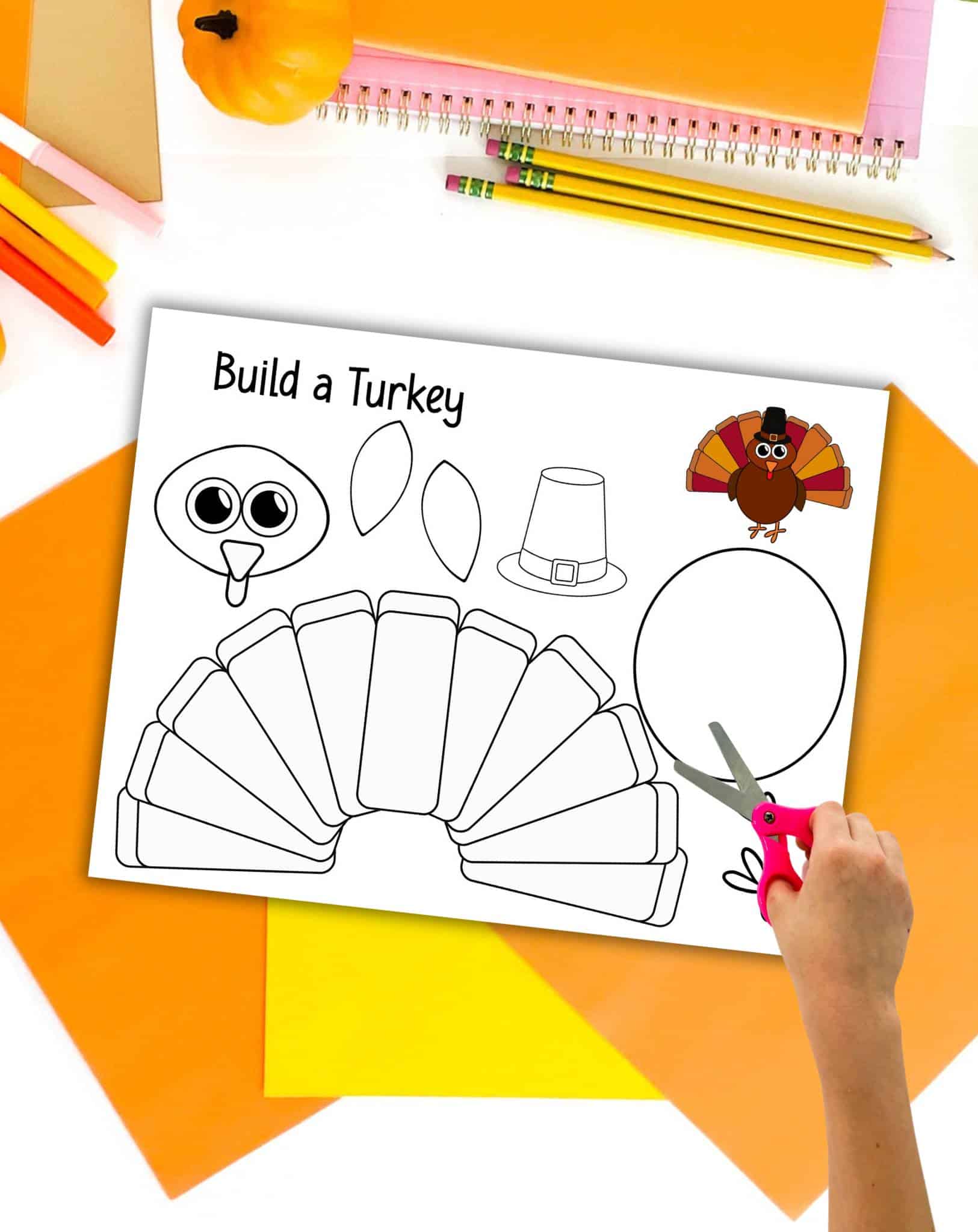 A preview of a printed build a turkey craft page with a child's hand with scissors. The page is shown on top of orange and yellow paper with fall colored craft supplies.