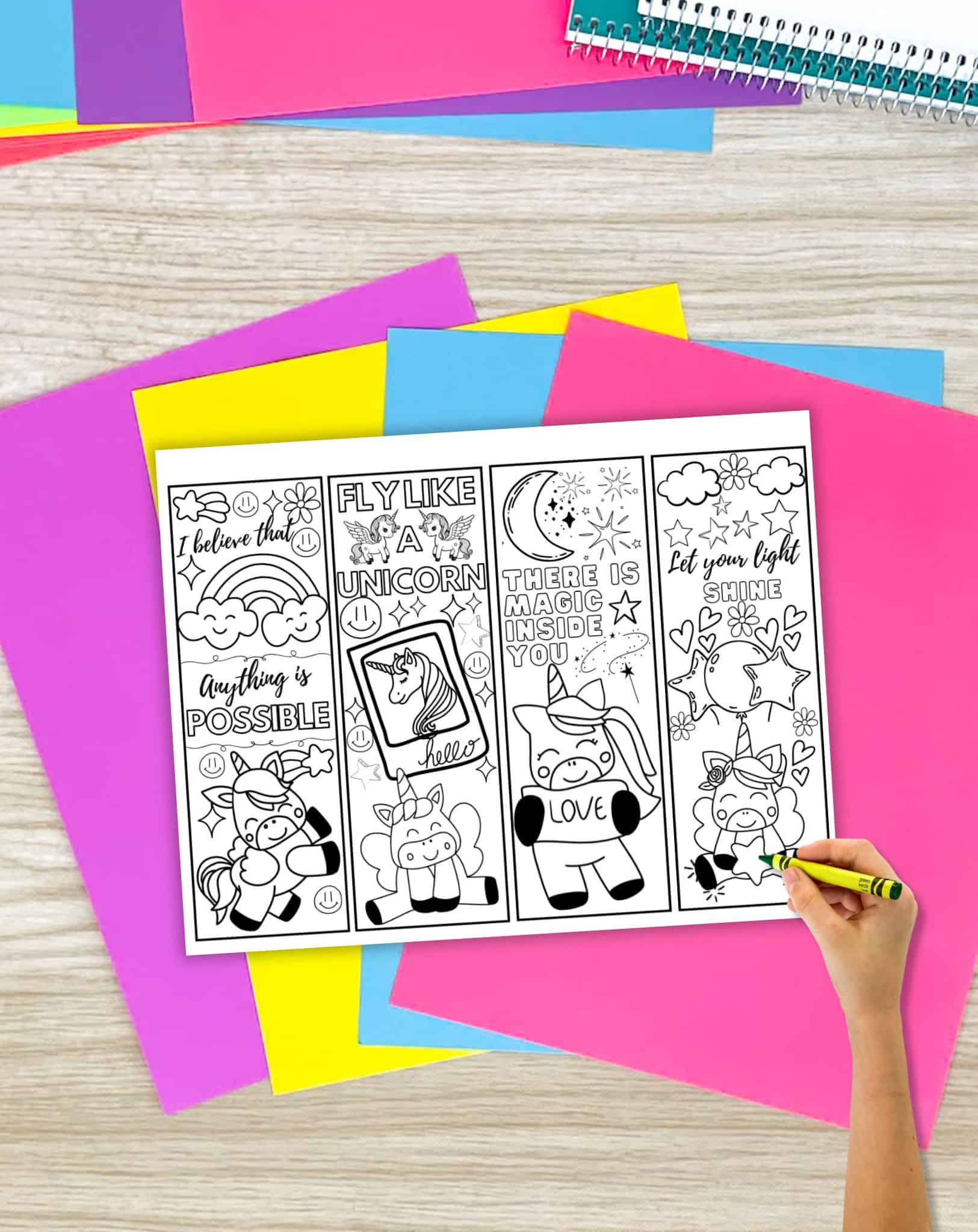 A page with four unicorn bookmark coloring pages on a desk with colorful papers and school supplies. A child's hand with a green crayon is ready to start coloring