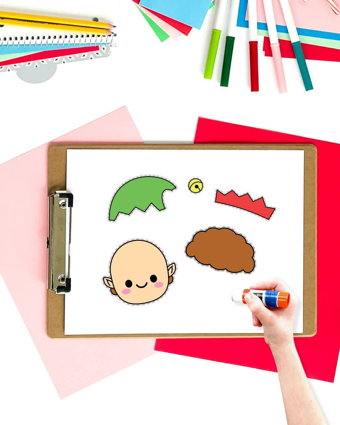 Pieces for an elf cut and paste craft on a clipboard with a child's hand holding a glue stick