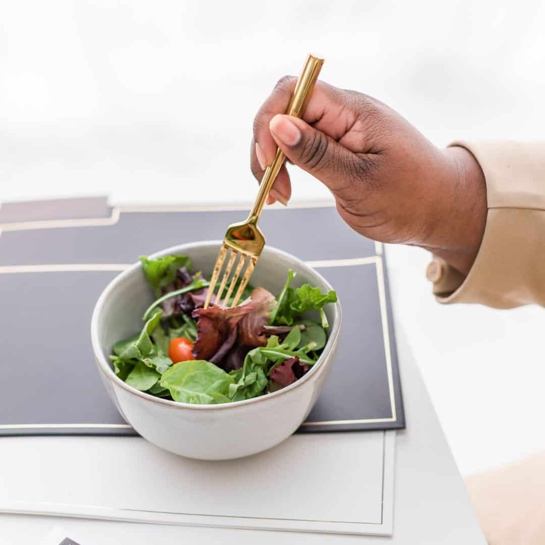 A woman's hand eating a bowl of salad which is resting on a desk