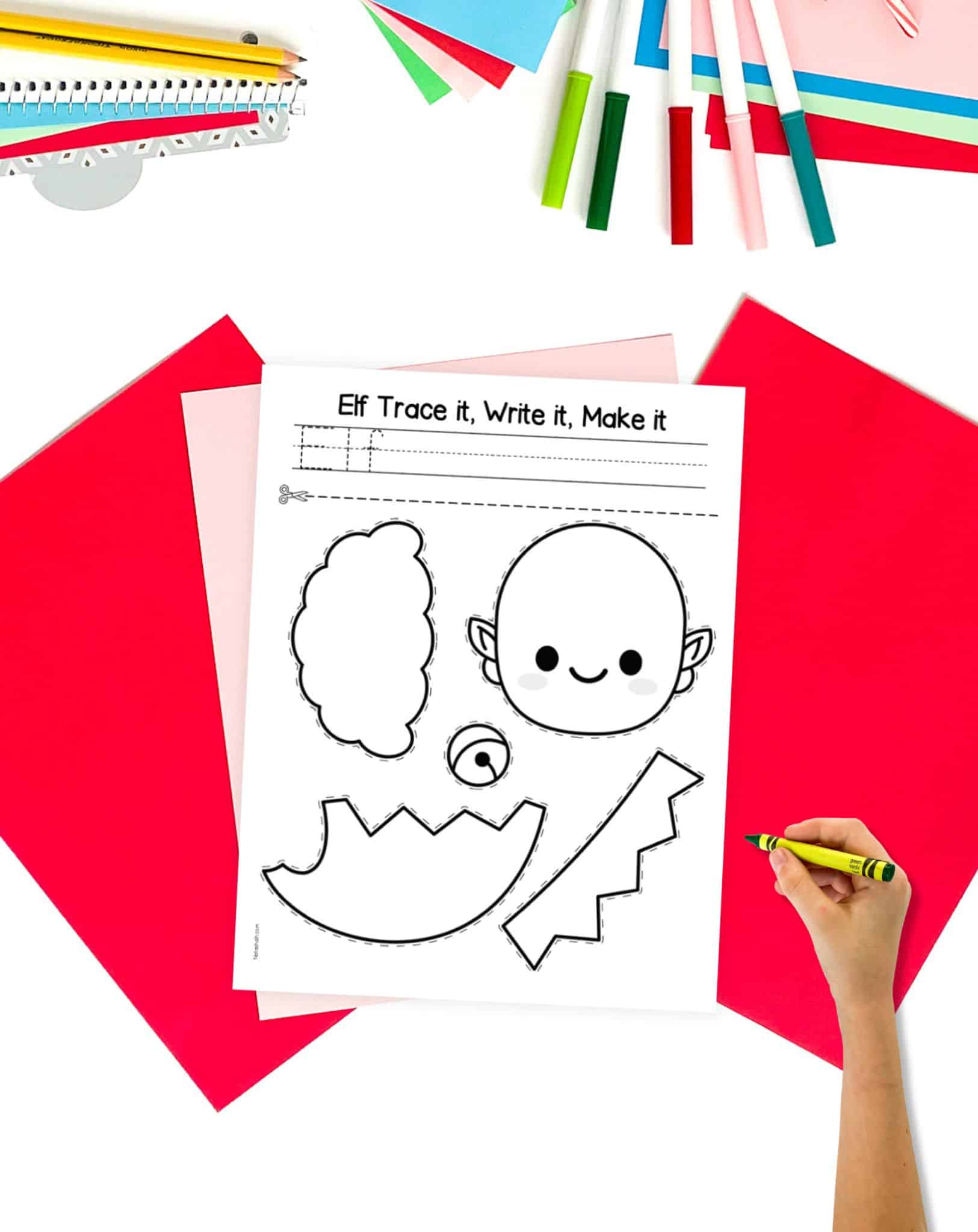 A black and white elf cut and paste craft on top of red and pink paper with a child's hand holding ag green crayon