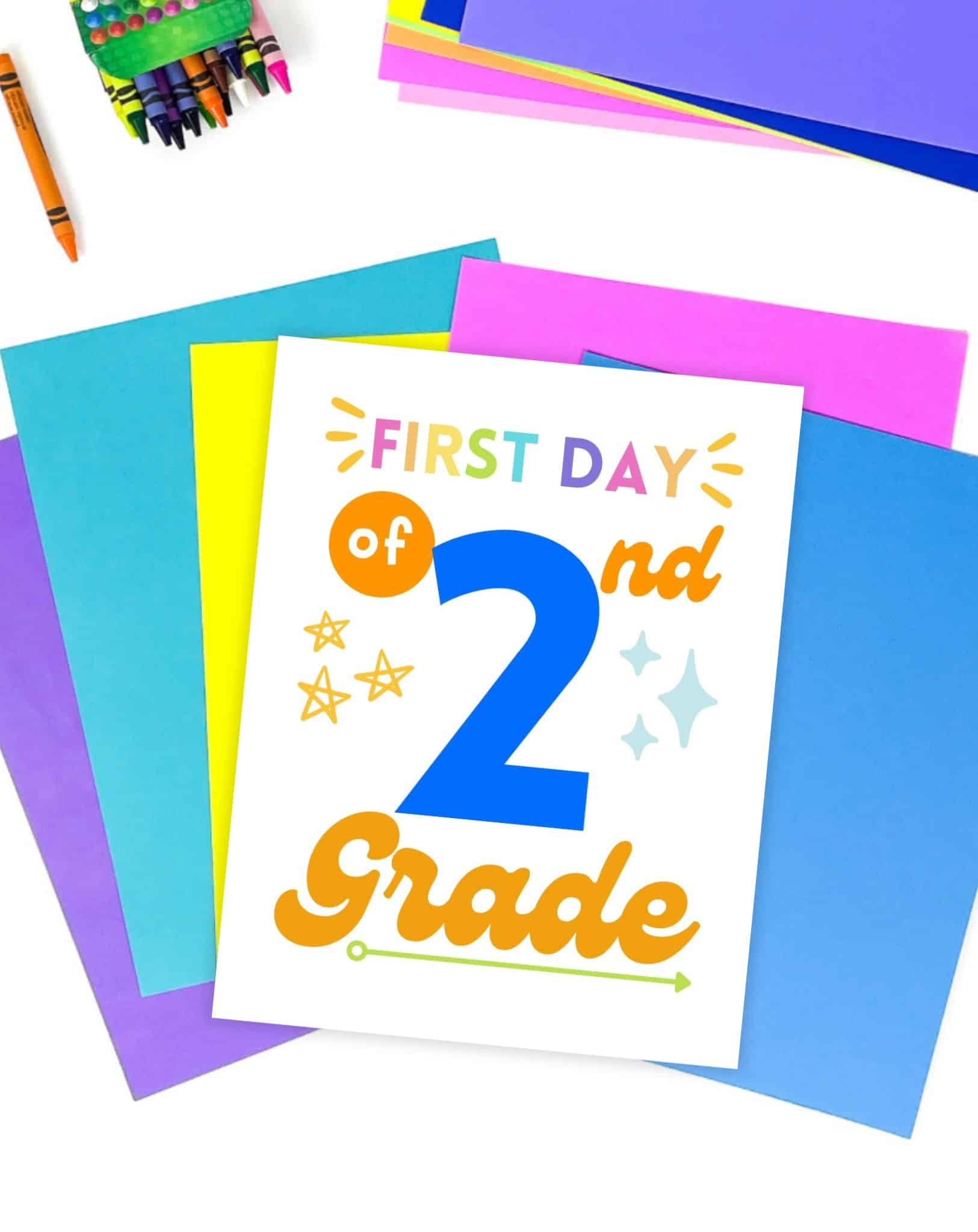 A first day of second grade sign on a desk with colorful papers and desk supplies