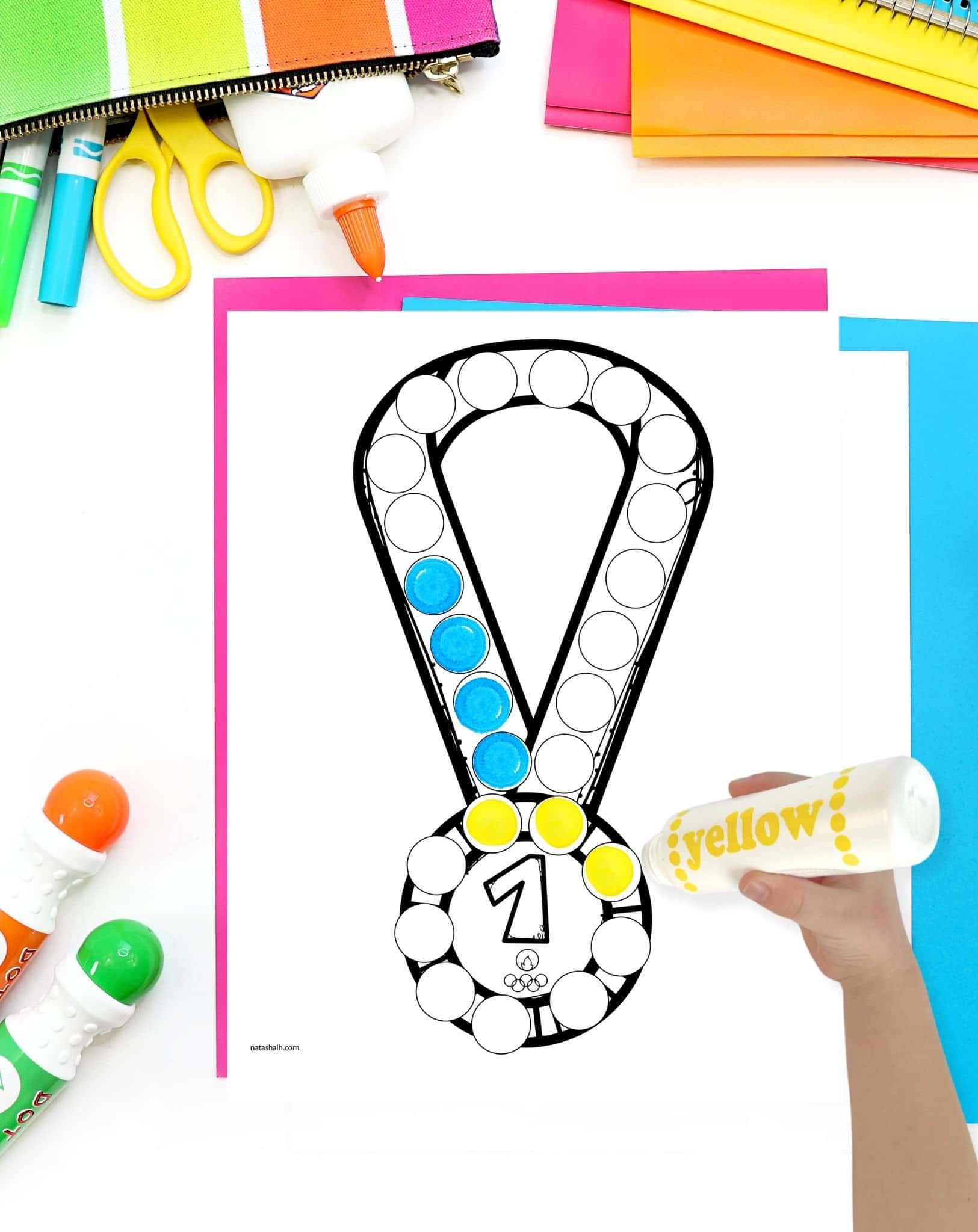 A top down view of a gold medal dot marker coloring page on a desk with brightly colored school supplies