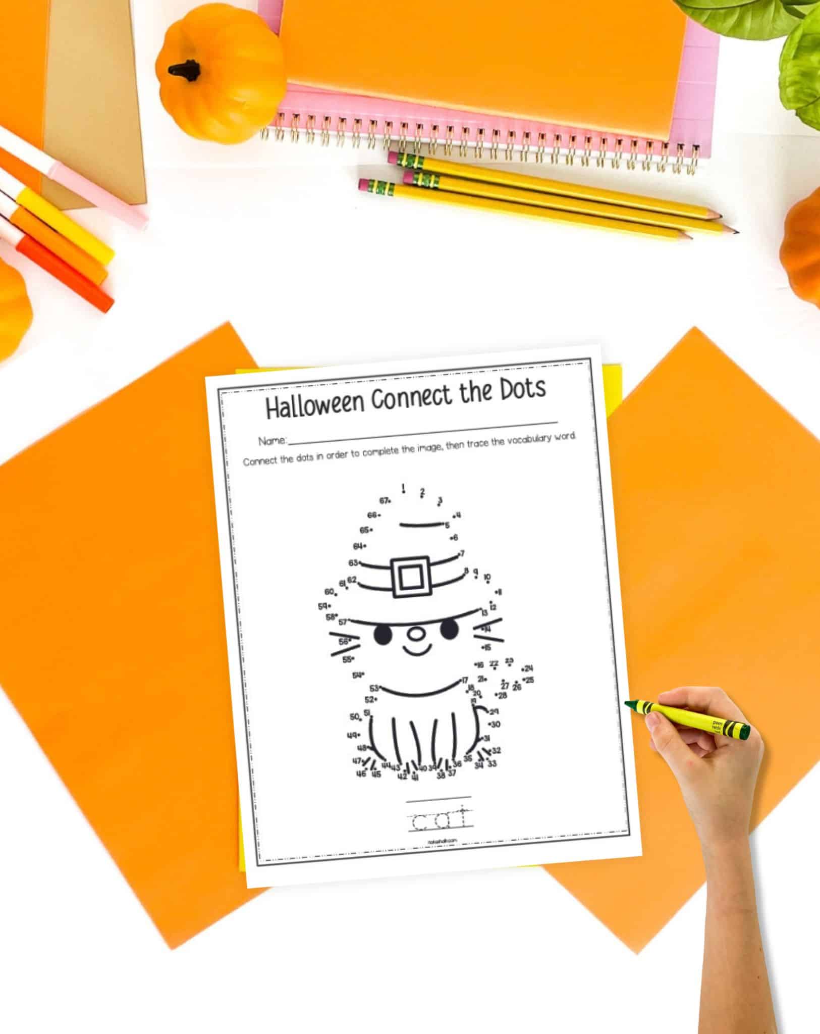 A Halloween cat connect the dots page with a child's hand holding a green crayon. The page is on a desk with fall colored papers and school supplies.