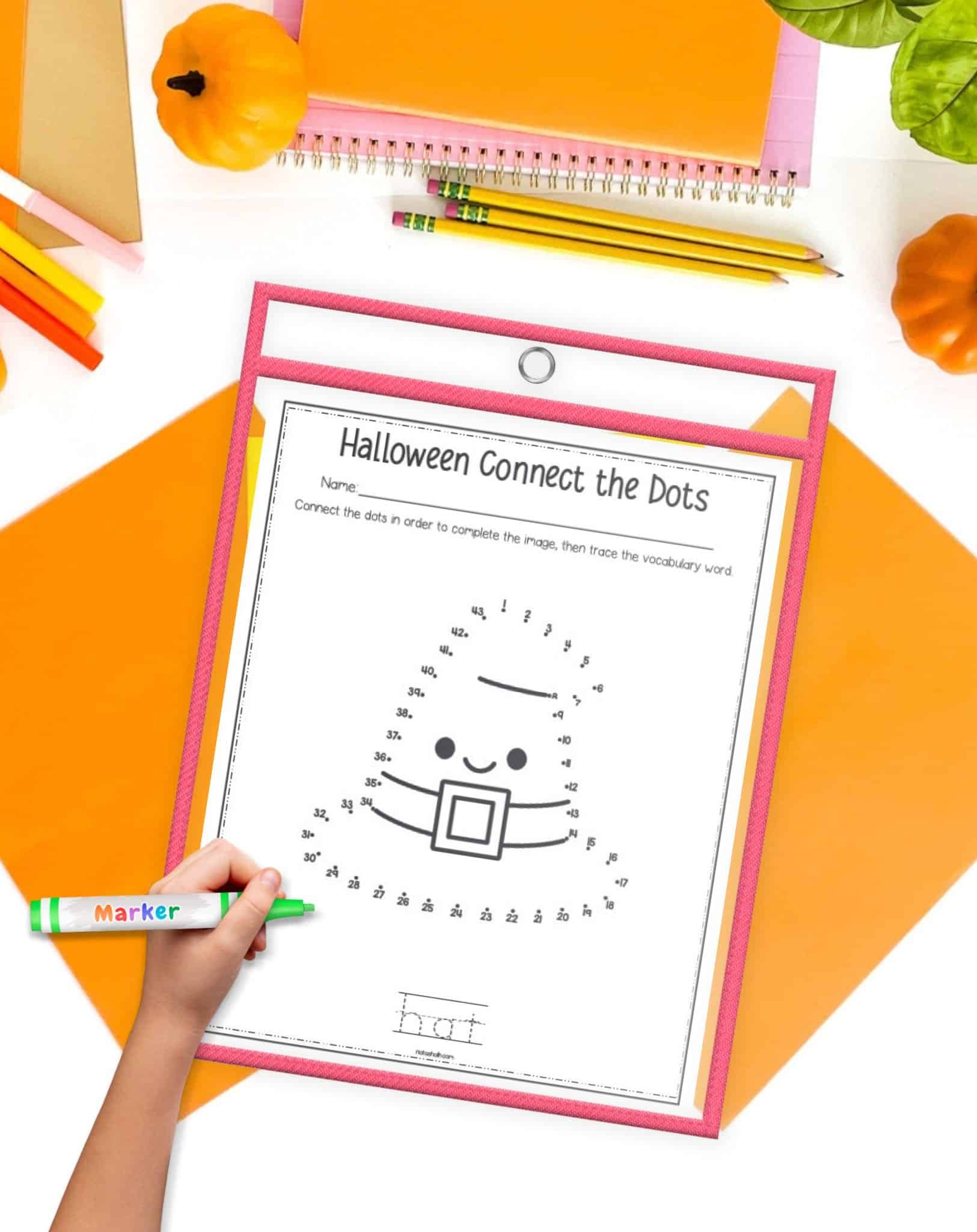 A Halloween witch hat connect the dots page in a dry erase pocket with a child's hand holding a green marker. The page is on a desk with fall colored papers and school supplies.