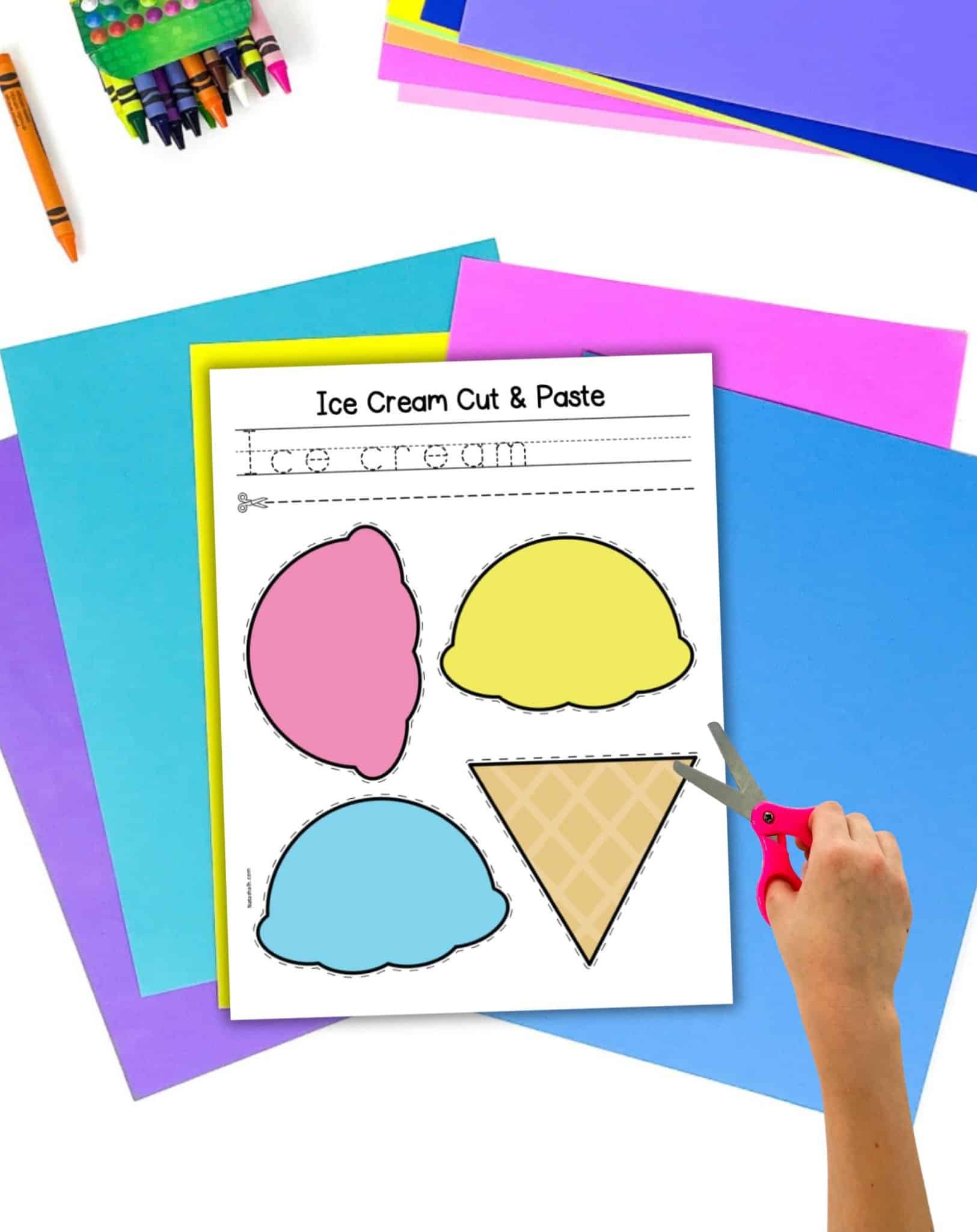 A top down view of colored paper, crayons, a child's hand with a pair of scissors, and a cut and paste ice cream craft.