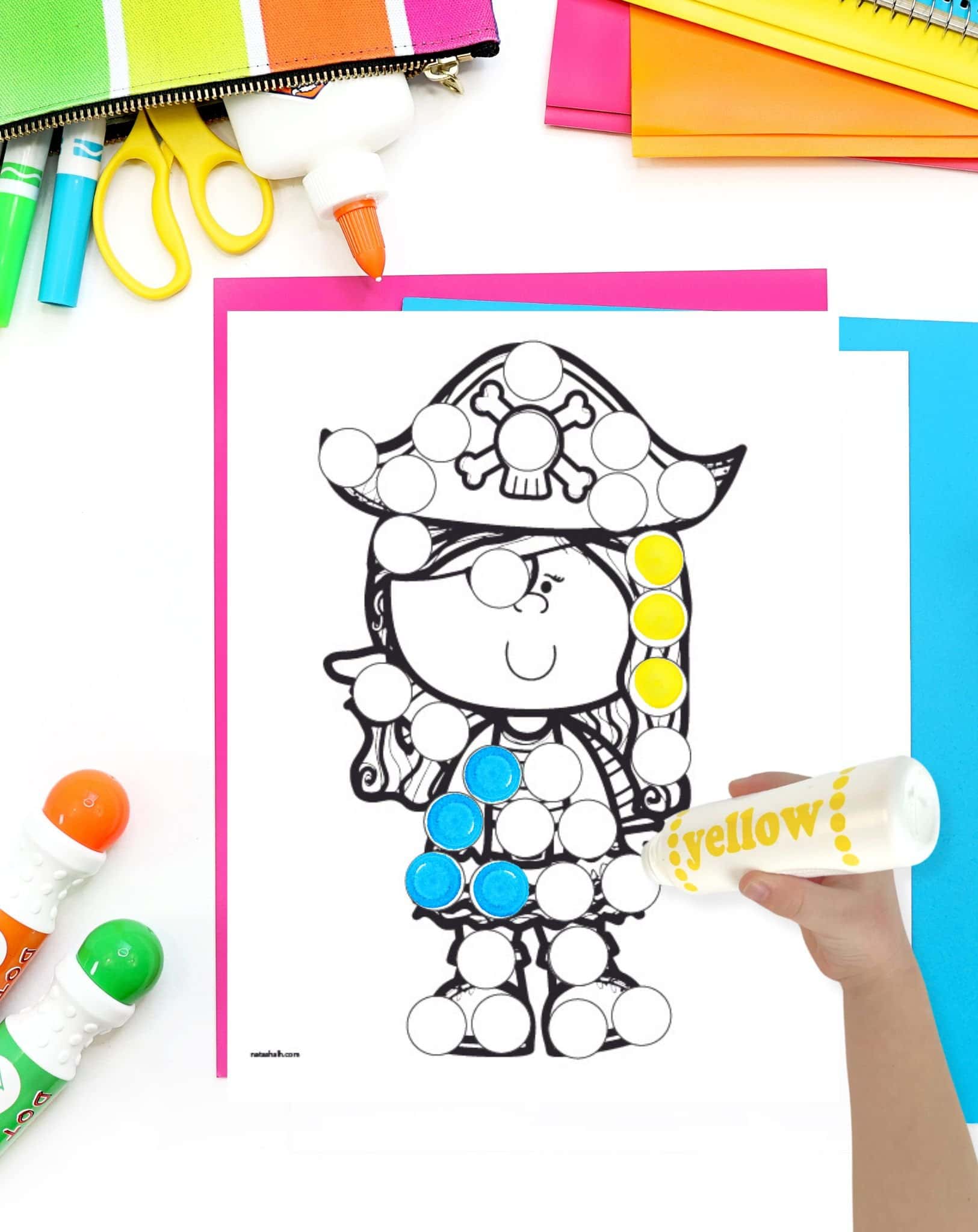 A pirate girl dot marker page on a desk with colorful school supplies. A hand holding a yellow dot marker is visible in the foreground