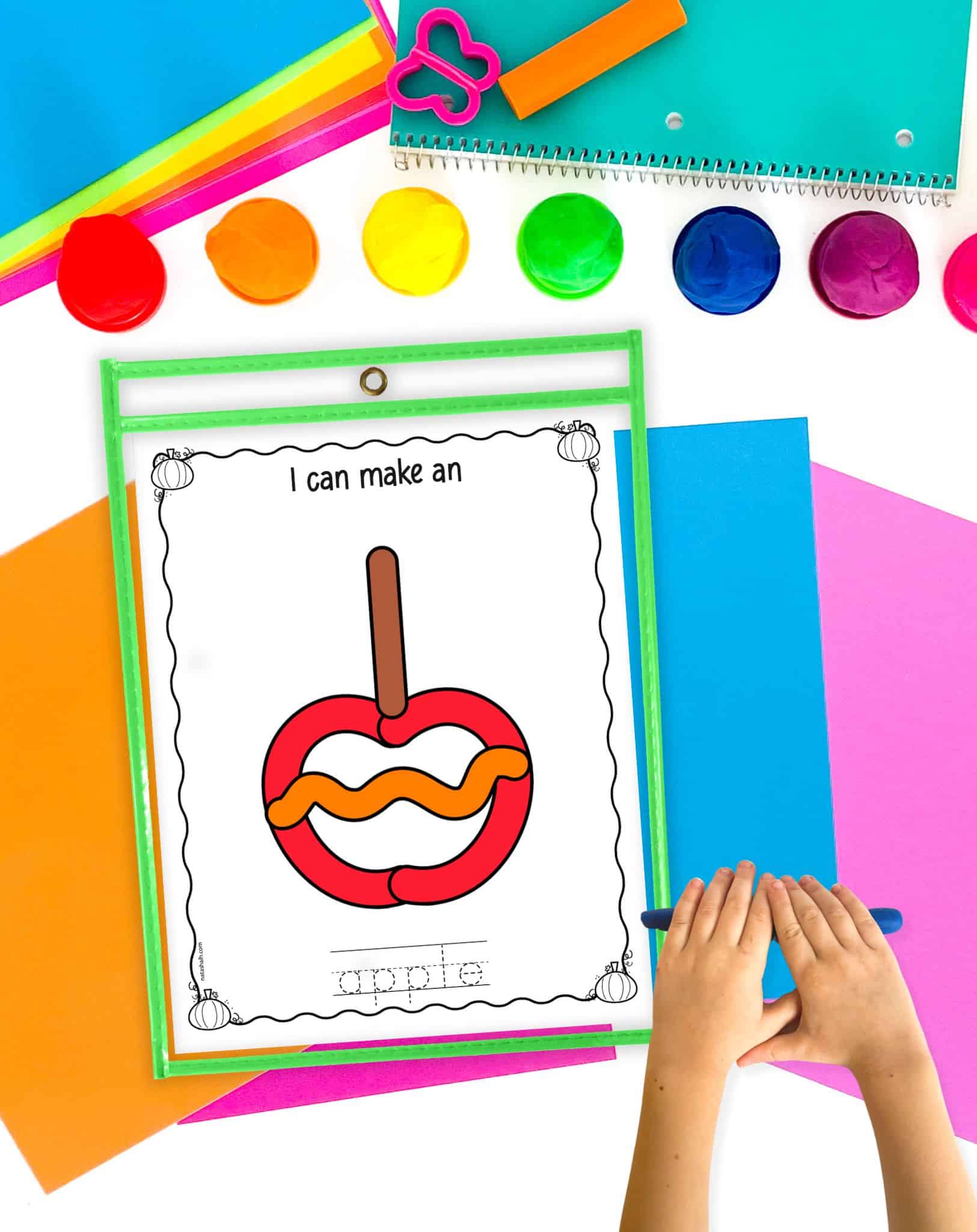 A candy apple play dough mat on a table with colorful papers and a child's hands rolling out a blue play dough snake