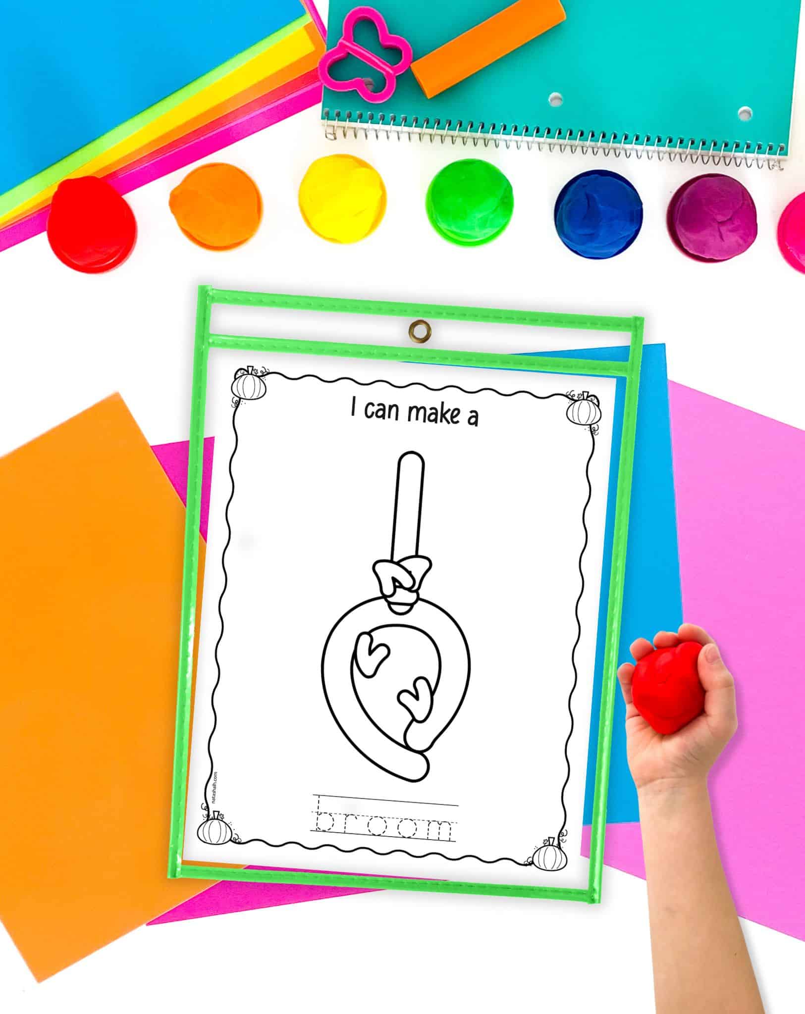 A broom themed play dough mat on a desk with colorful papers and a child's hadn't holding a ball of play dough