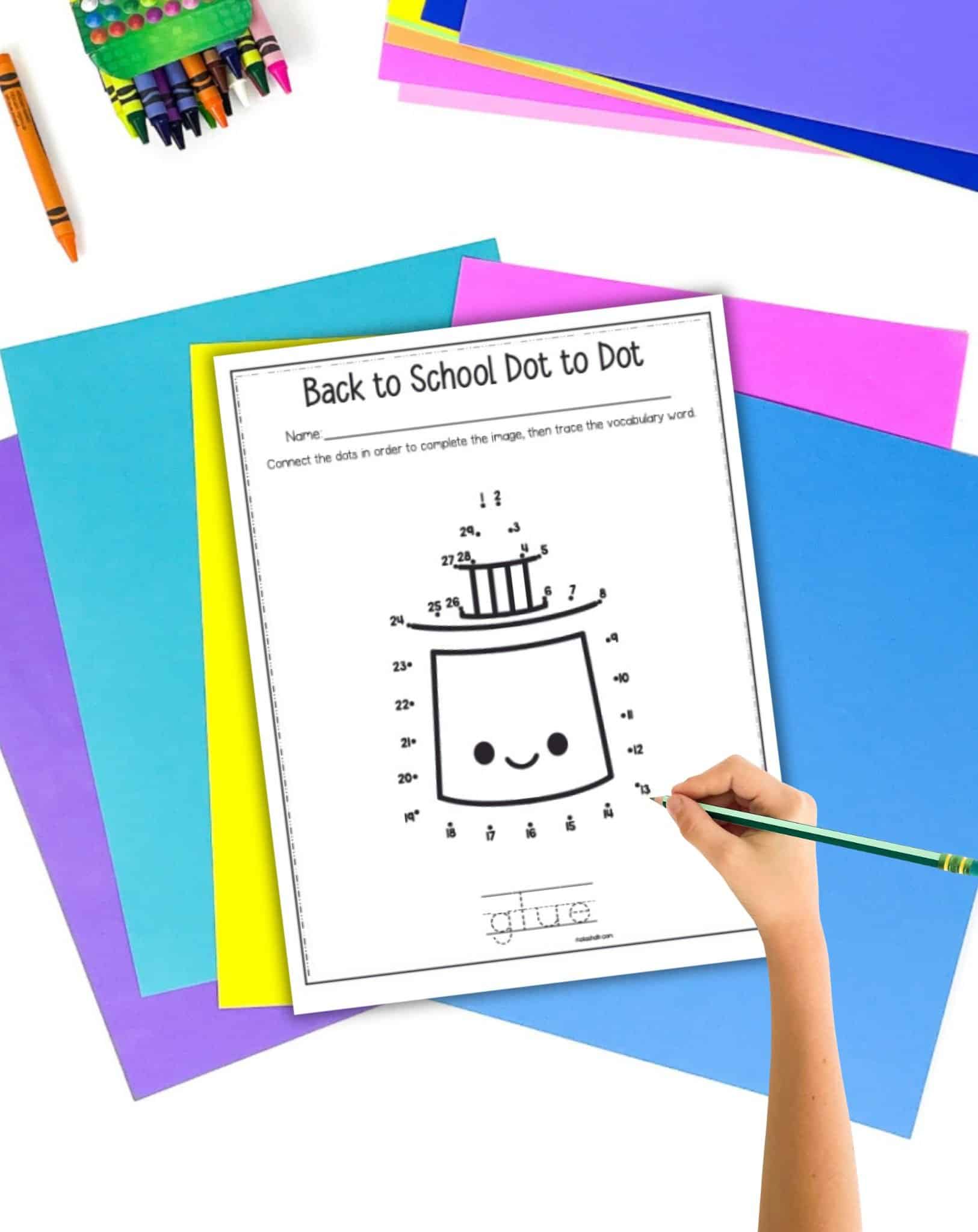 A top down photo of a child's hand holding a pencil over a back to school themed connect the dots page showing a glue bottle