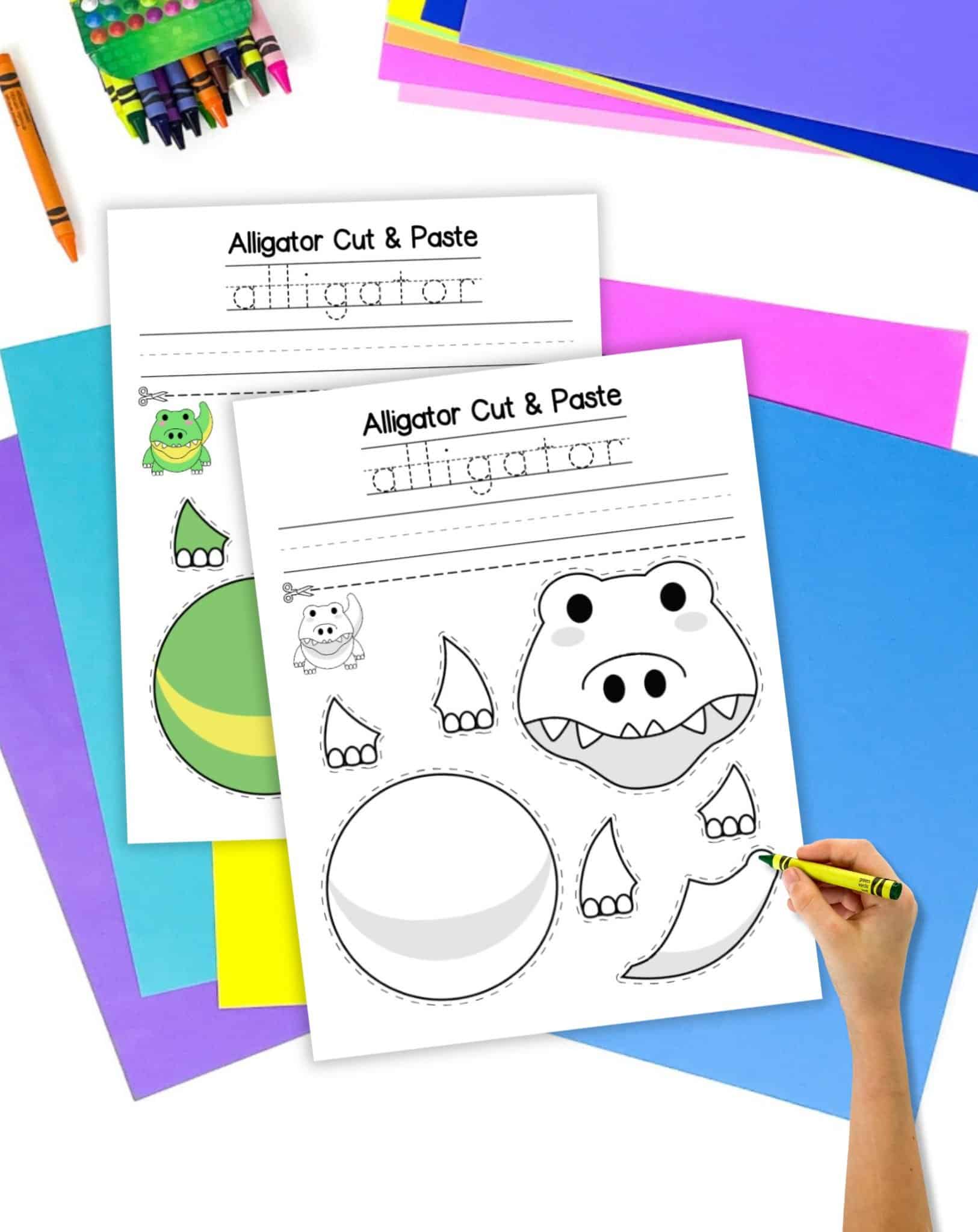 A top down view of a two printable cut and paste alligator crafts on a desk with colorful papers and crayons. One is color and the other black and white.