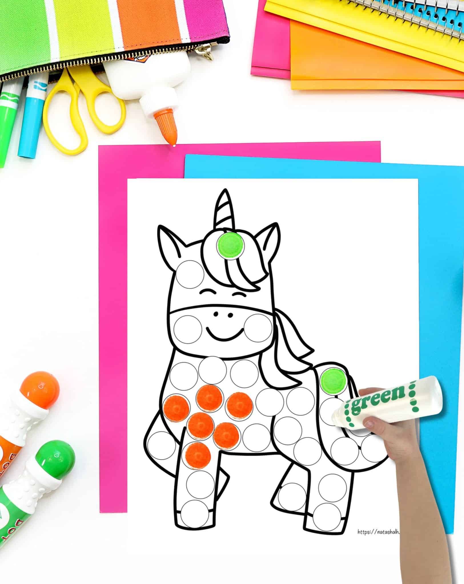 A top down photo of a unicorn dot marker coloring page with bright school supplies on a desk