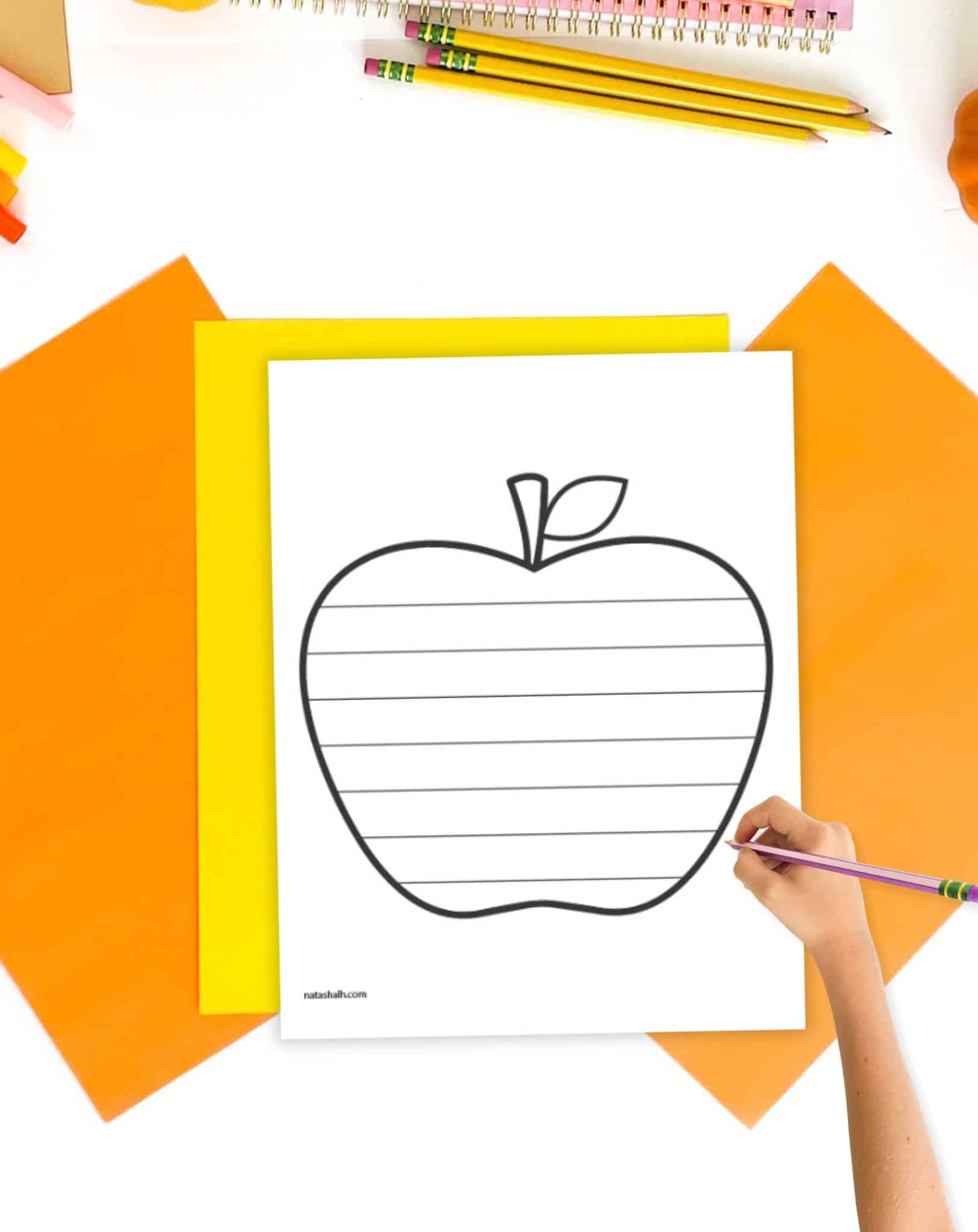 A themed writing paper shaped like an apple on a desk with fall colored papers. A hand holding a pencil is visible in the foreground 